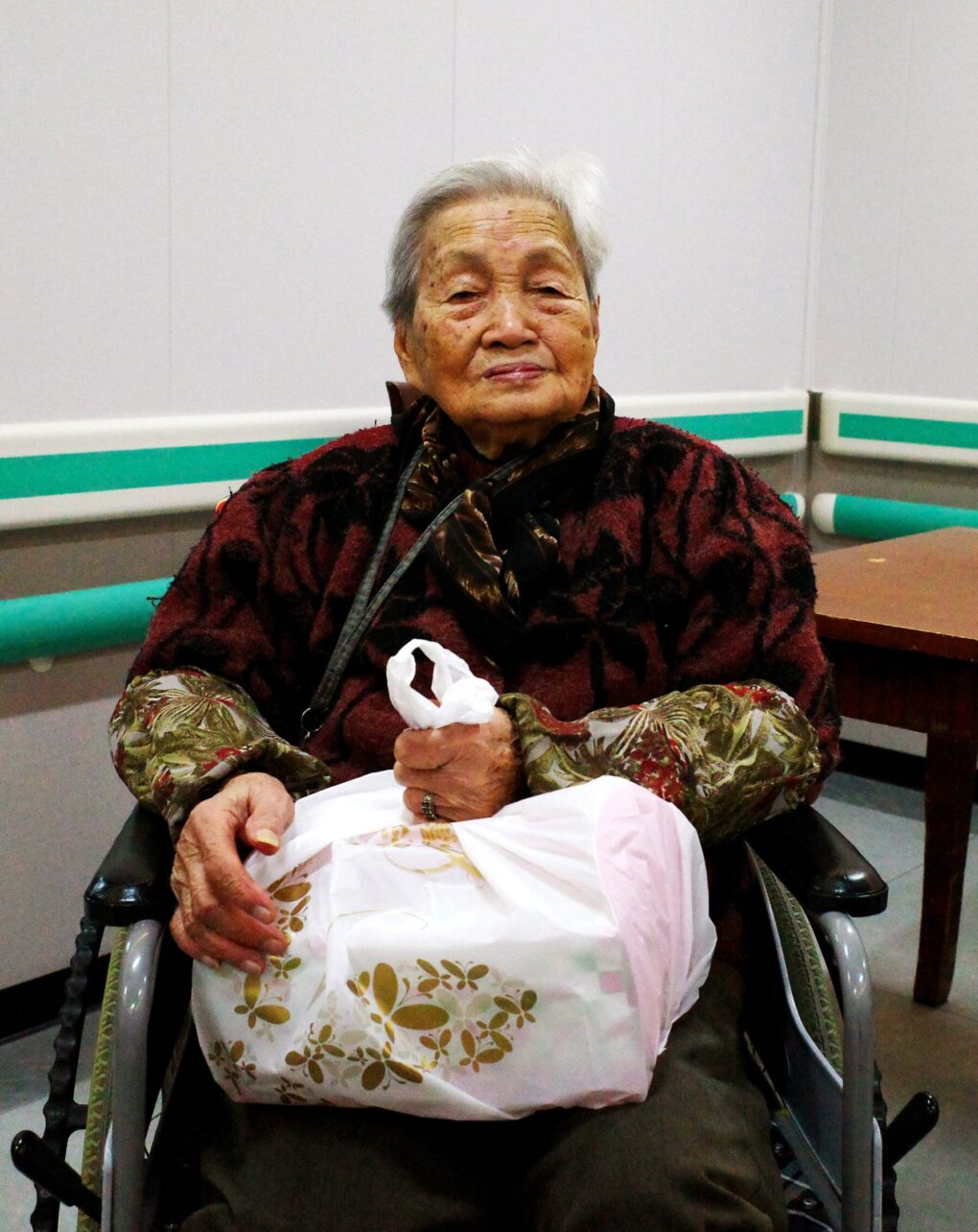 An elderly Chinese woman with white hair and age spots is sitting in a wheelchair looking directly at the camera.