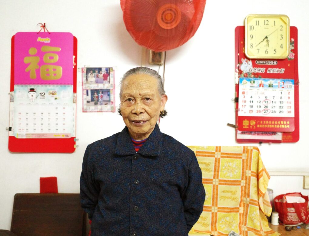 An older Chinese woman is standing between two calendars, smiling, apparently in her home.