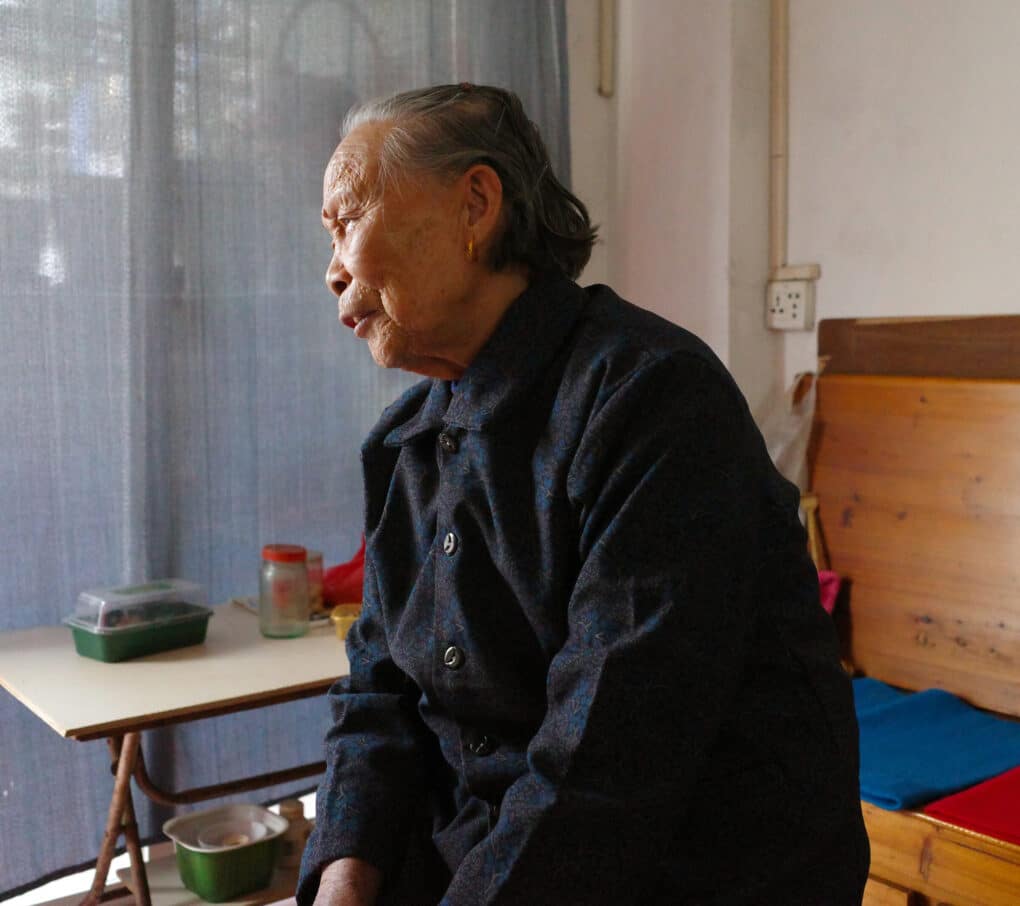 An elderly Chinese woman with gray brushed-back hair is speaking. She's wearing a black and blue buttoned up jacket.