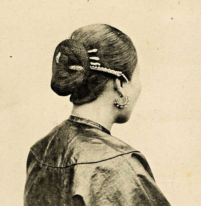 Black and white engraving of a woman's hairstyle, pictured from behind. A bun secured with decorative sticks and netted.