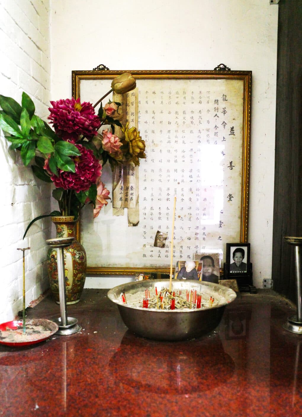 Photo of countertop with bowl with incense stubs, magenta dahlias and pink roses, and a large page of Chinese names in calligraphy, in a gold frame.