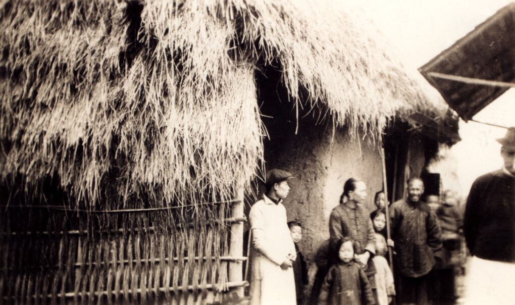 Very old photo of several people and children standing in front of a structure with a thatched roof made of grasses.