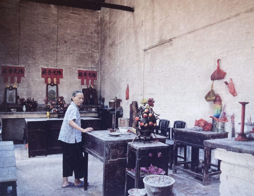 A woman stands at a dark wood table, with three altars to her side, and offerings of fruit and incense nearby.