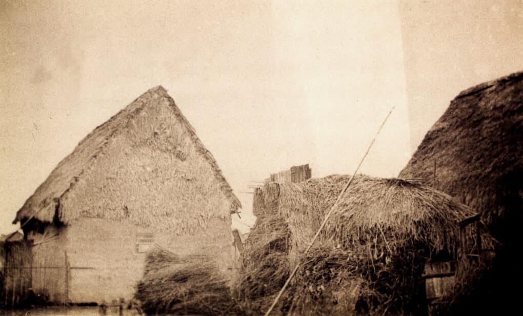 Very old black and white photo of building with pitched roof, made out of mud and grasses.