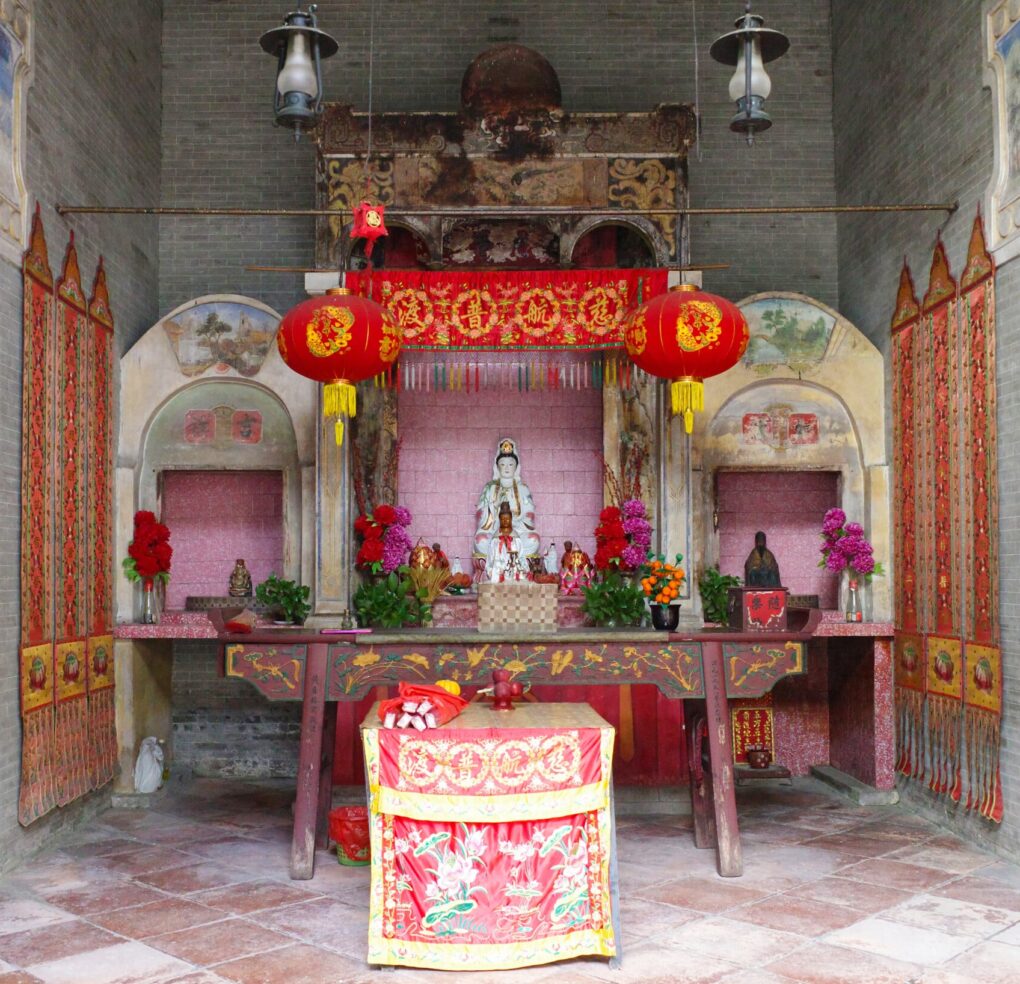 Brick room with high ceiling and elaborate alter with statue at center and pink and red flowers, silk banners, lanterns, and various offerings.