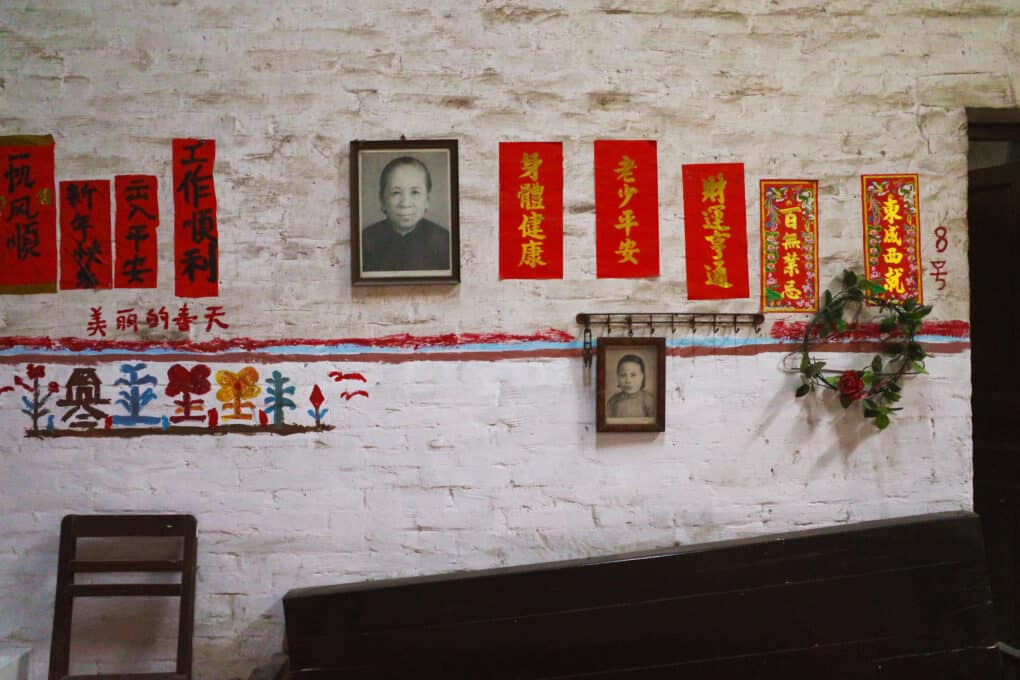 White wall with portrait photo of Chinese woman in center, and many vertical rectangles of red paper with black lettering hanging on both sides.