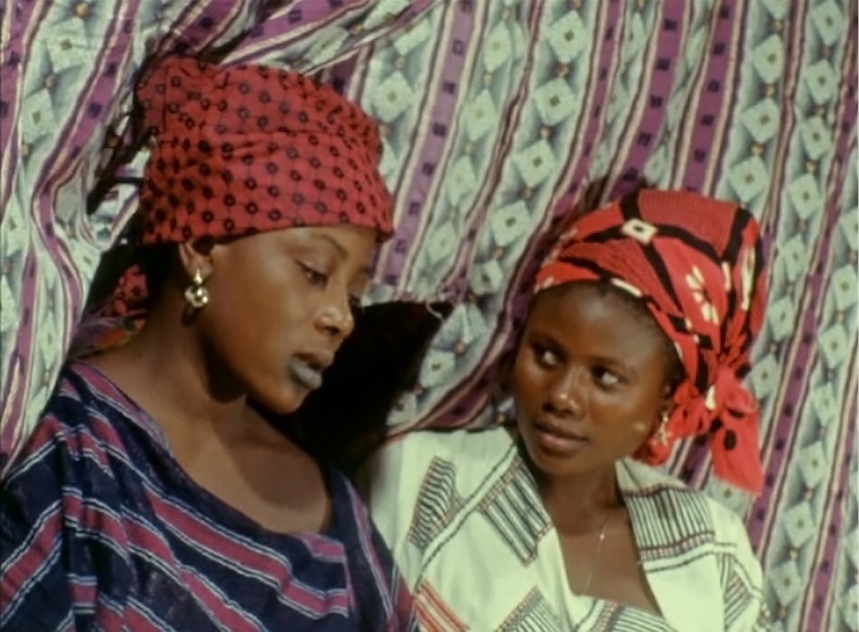 Color film still of two young Black women in red-and-black headwraps, against a pink-and-white-striped cloth backdrop.