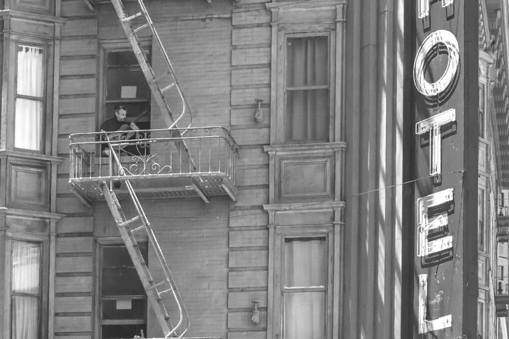 A man is playing guitar, perched out his window, with his feet resting on the fire escape. The Ambassador sign is visible to the right.