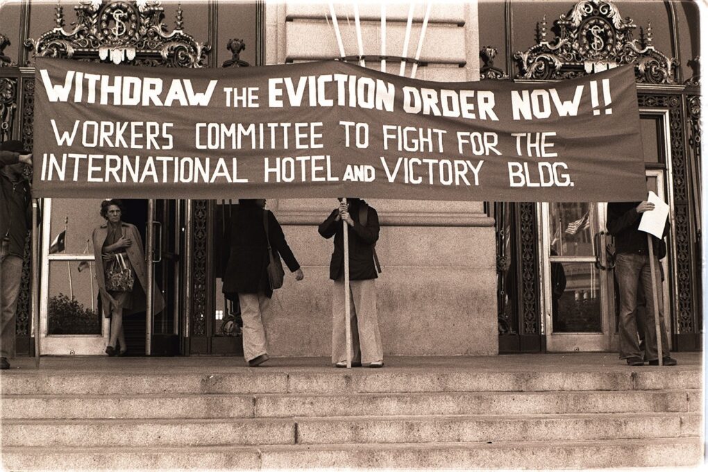 Several people are holding a very large banner in front of a building. The banner says, "Withdraw the Eviction Order Now!! Workers Committee to Fight for the International Hotel and Victory Building."