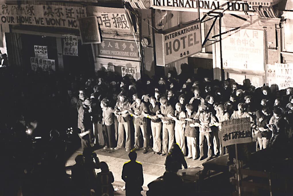 High angle view looking down on about a block of about fifty protestors standing with linked arms beneath sign for the International Hotel.
