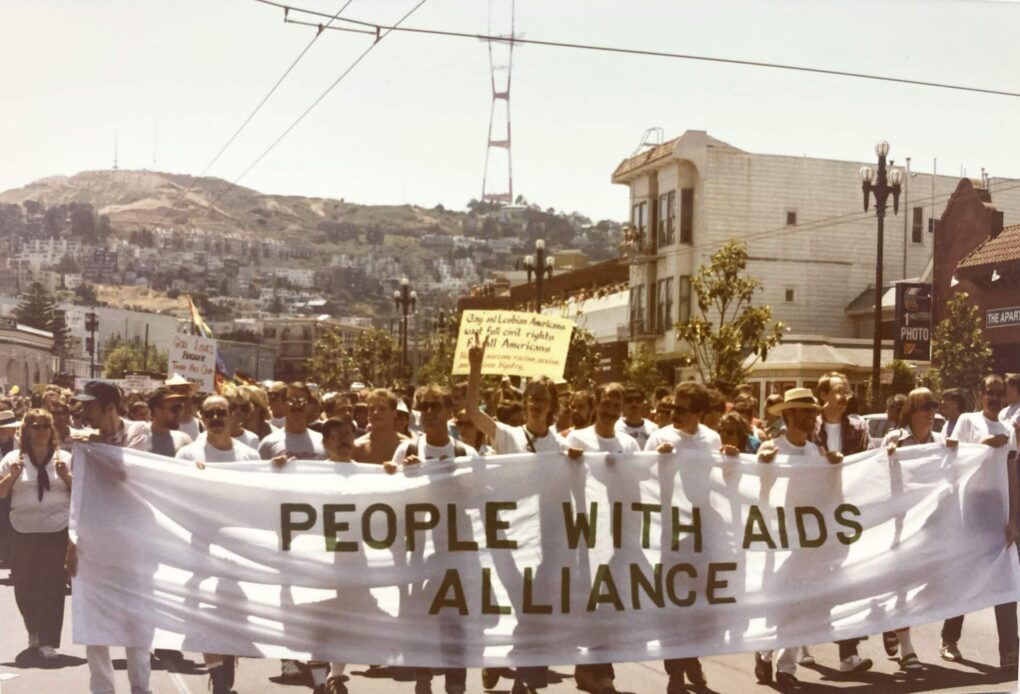 Front of public march, with many people holding banner that says "People with AIDS Alliance."