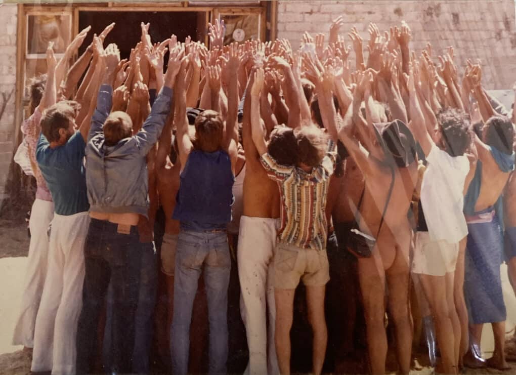 A group of men tightly clustered are all raising their arms and hands in unison. Some men are only partially clothed, no faces visible.
