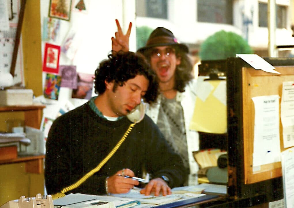 A man with curly brown hair is cradling the phone on his shoulder and taking notes, while someone behind him makes the peace sign, goofing off.