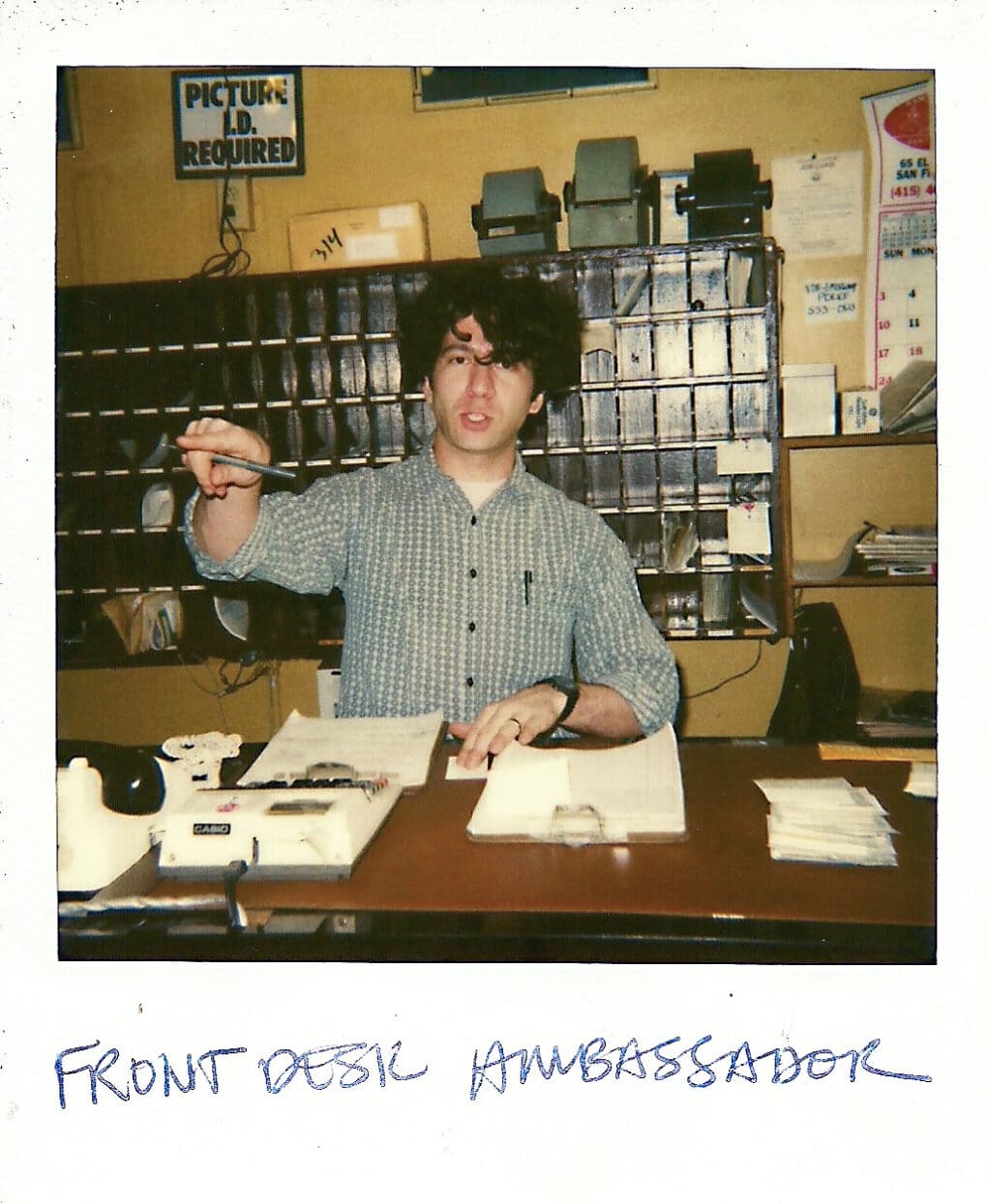 A Polaroid snapshot of man in his early 30s wearing a button-up shirt, with curly dark hair, standing behind a counter with a clipboard, adding machine, and various papers. He appears to be pointing, as if giving directions.