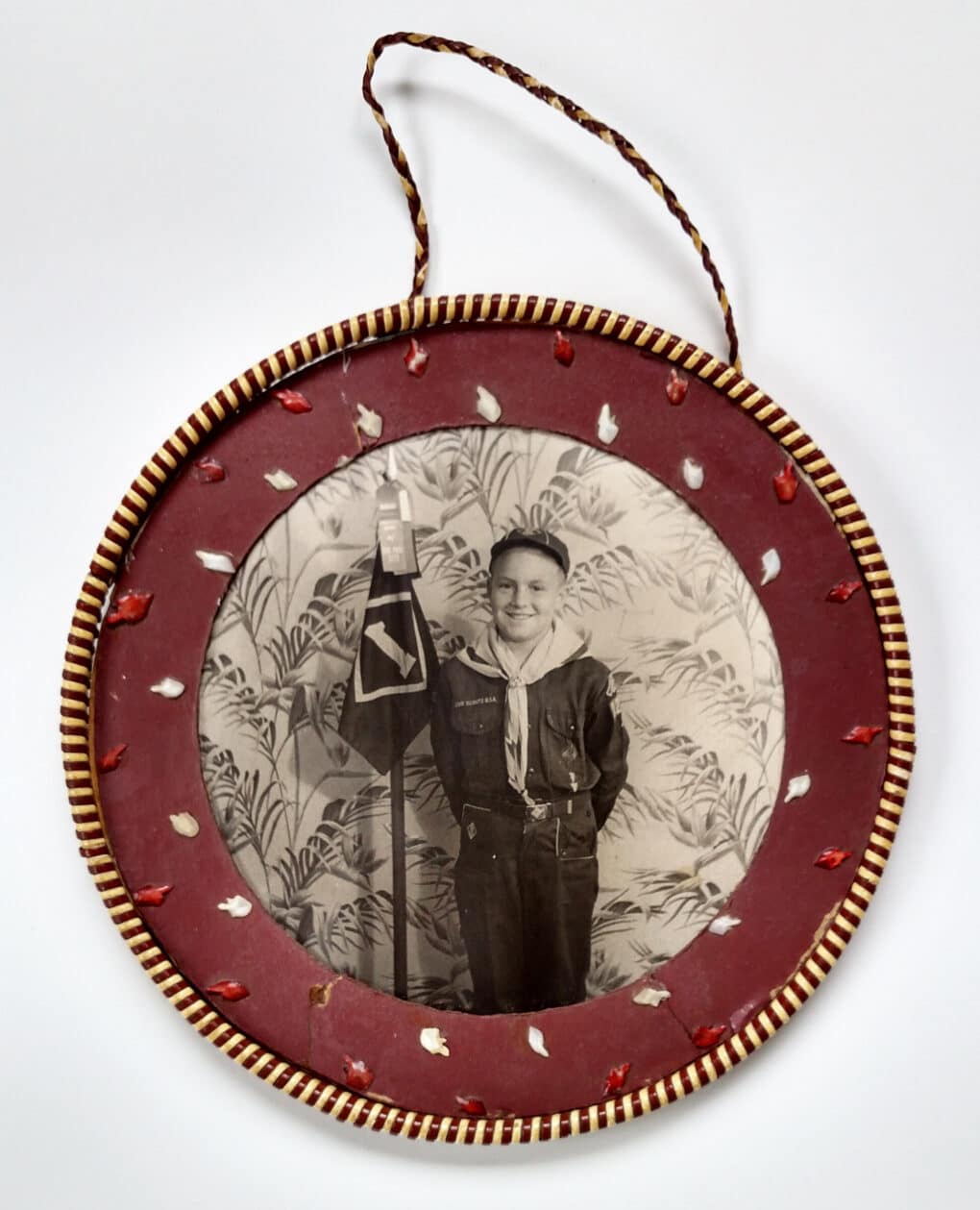 Circle pendant photo of young boy in Boy Scout uniform smiling broadly and holding pole with ribbons attached, as if he's won an award. 