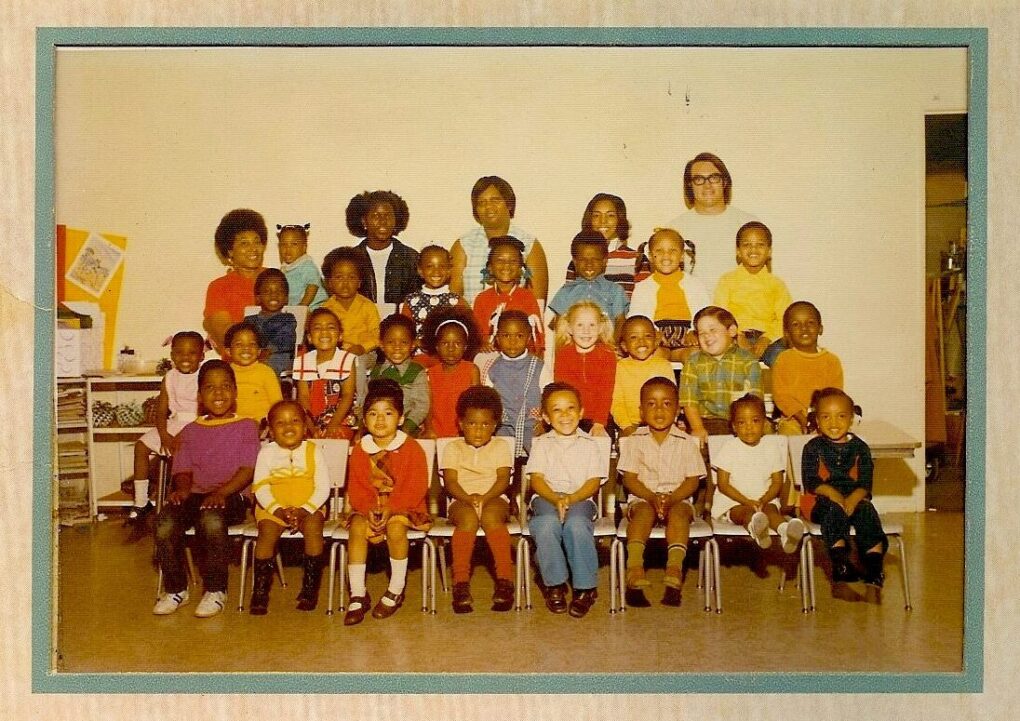 Classic class picture of small children with their teacher. Majority of the children are black, front row is seated, and there are 5 adults at the back, 4 black women and 1 white man with longish hair.