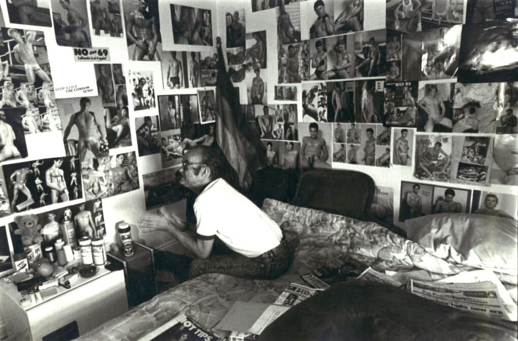 A man is sitting on a bed, and all around him the walls are papered with cutout of naked men. 