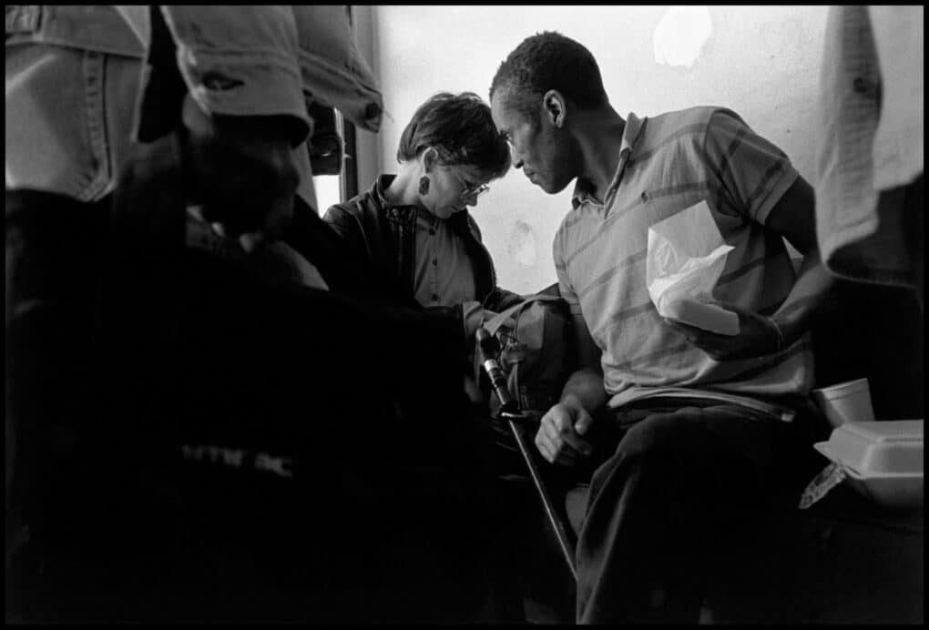 A black man is seated, listening attentively to a woman with short hair beside him, who seems to be consulting notes. There are other people in the frame, their faces not visible.