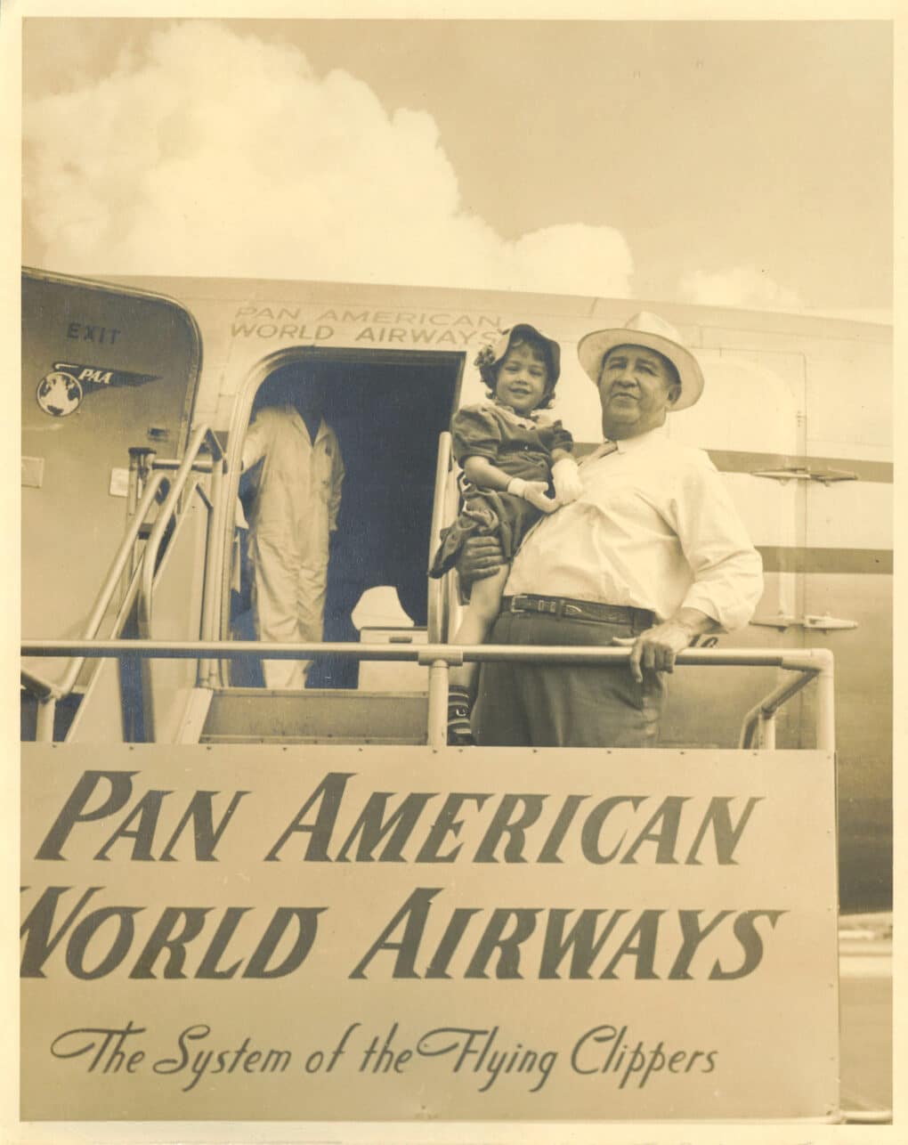 Black-and-white photograph of small girl with bow in her hair, held in the arms of a large man in a white shirt and Panama hat; they stand on the jetway stairs of a small airplane, with a sign in the foreground reading "Pan American World Airways: The System of the Flying Clippers."