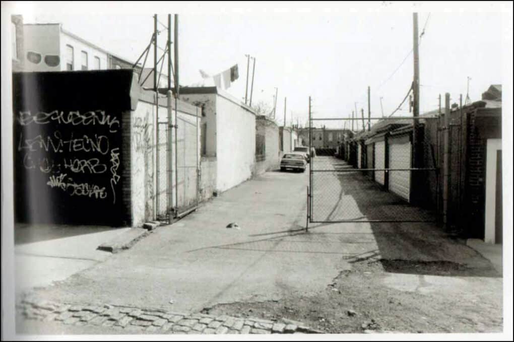Black-and-white photograph of nondescript urban alleway with open chainlink gate.