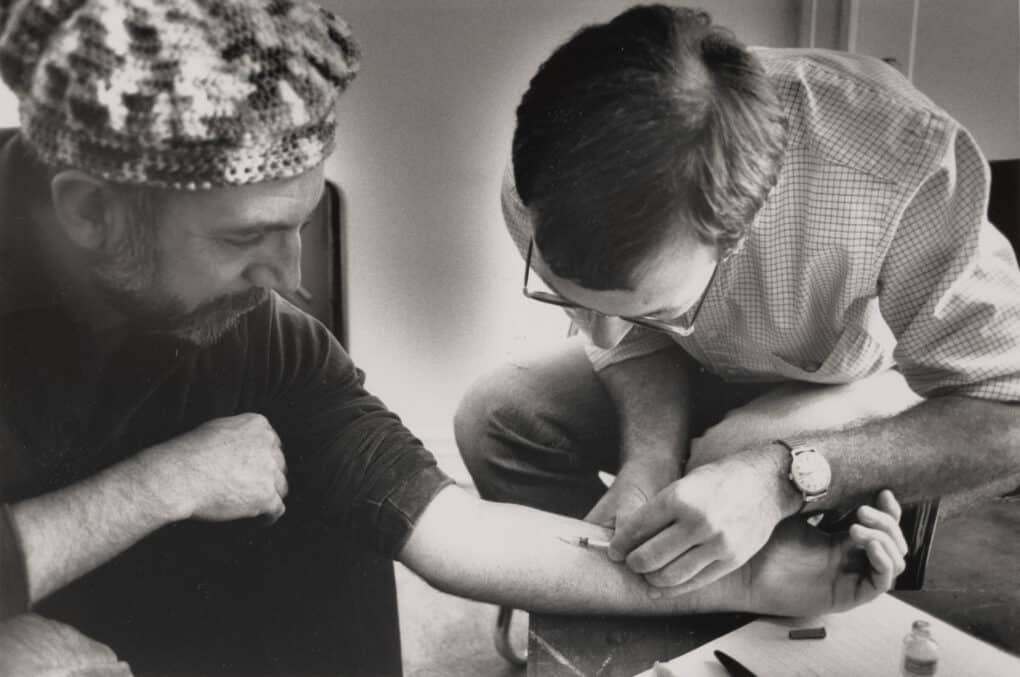 One man leans over another man's arm and inserts a hypodermic needle into his forearm. 