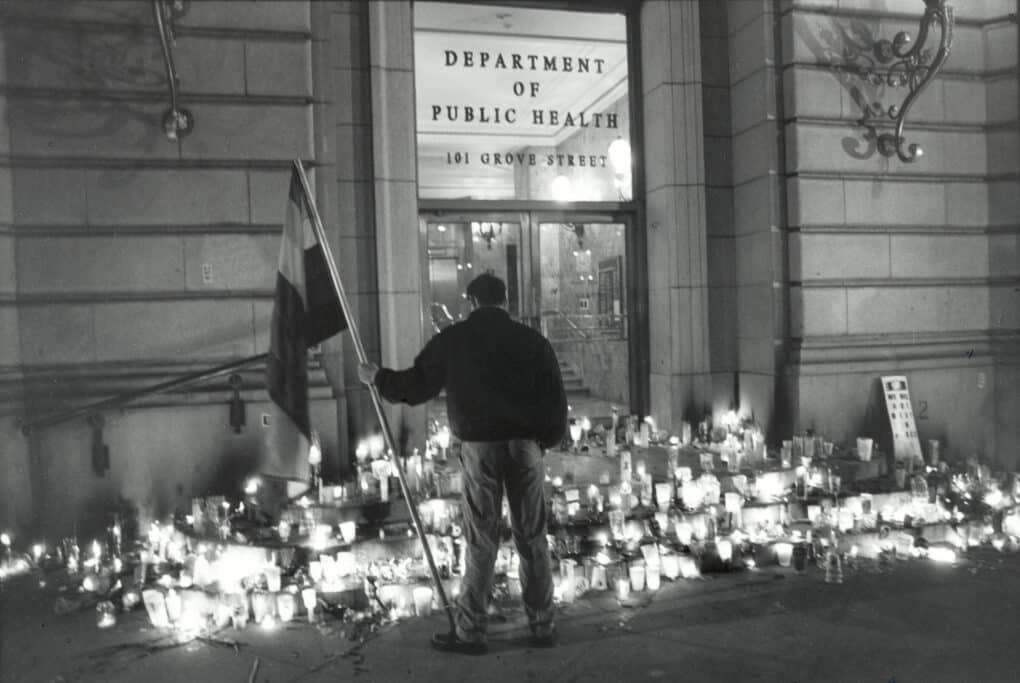 Dozens of candles on the steps to a building door marked "Department of Public Health." One man is standing with his back to the camera, looking down at the candles and holding a rainbow flag.