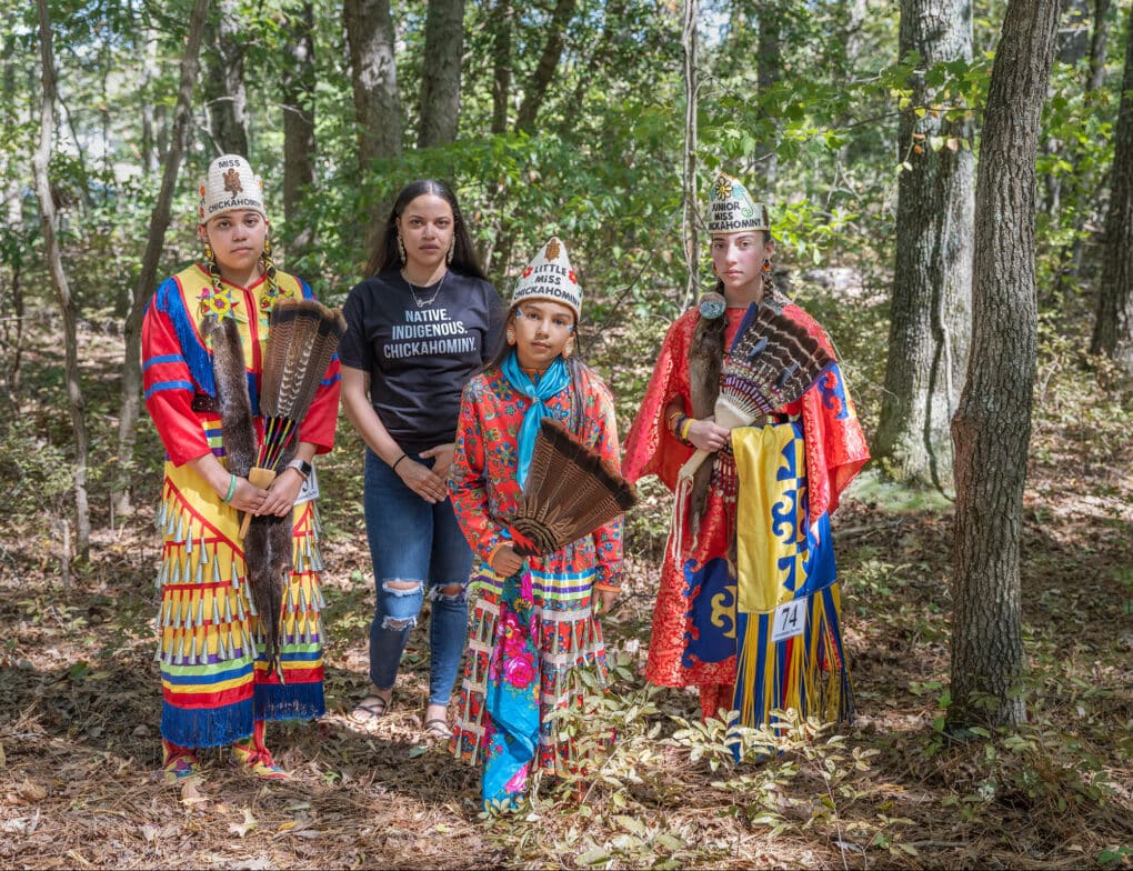 Color photograph of four young women standing in the woods. Three wear colorful skirts, shawls, and headdreses, and carry feather fans. The fourth wears a t-shirt that reads: NATIVE. INDIGENOUS. CHICKAHOMINY.