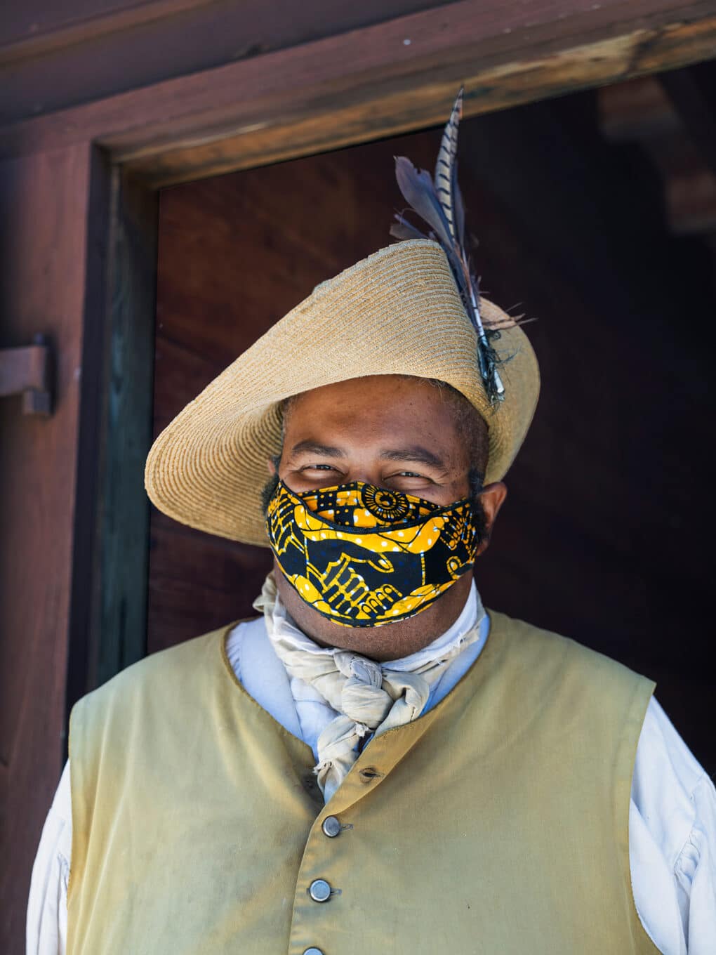Color portrait of heavyset black man wearing period dress and a kente-cloth mask, smiling with his eyes at the camera.