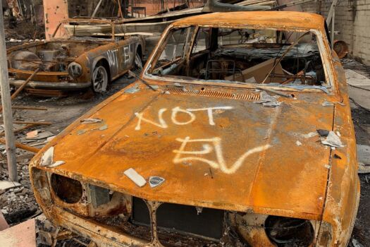 Color photograph of burned-out car, rust-red, with "NOT EV' spraypainted on the hood.