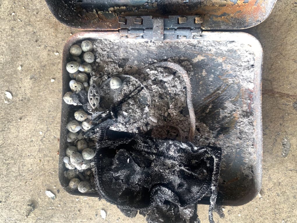 Color photograph of small burn-scarred metal box, filled with ash, along with some white beads and other bits of metal.