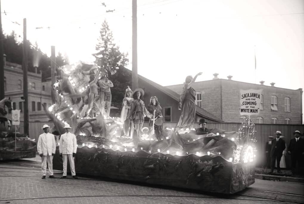 Black and white of parade float with figures standing on illuminated, decorated platform. Figures are dressed as Native Americans, fur trappers, etc. and in the front is the figures of Sacajawea, pointing. A sign says, "Sacajawea and the Coming of the White Man"