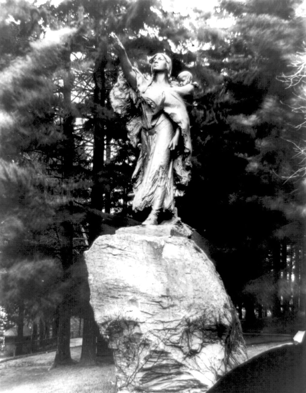 Old black and white very dramatic photo of a figurative sculpture, depicting a woman standing on a giant rock, with her hand strongly outstretched, and a child on her back. 