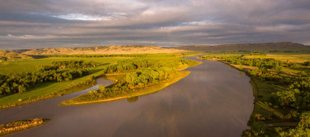 Pretty landscape photo of calm river surrounded by green fields, brush, and trees, with a very big sky. 