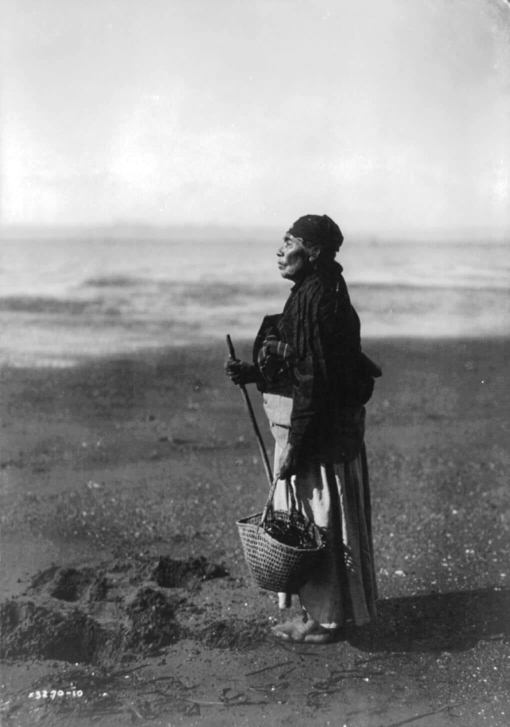 A Native woman is standing in profile, looking at the water. Her head is wrapped in a scarf, and she's carrying a basket and holding a staff. 