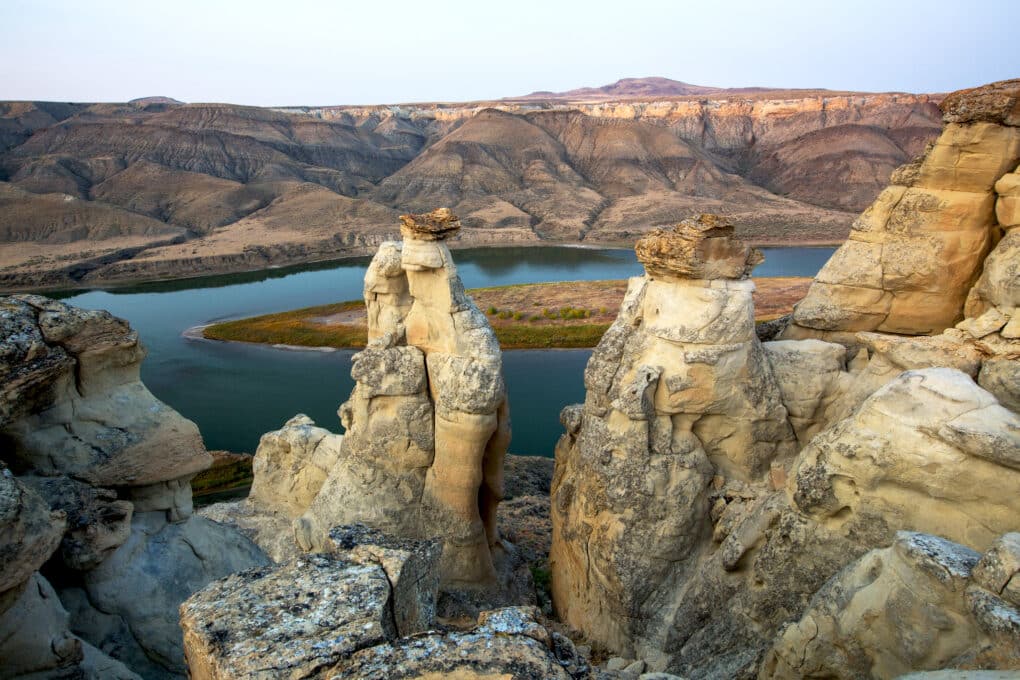 Dramatic landscape photo of strangely shaped rock croppings rising above blue water, "painted hills" in background. 