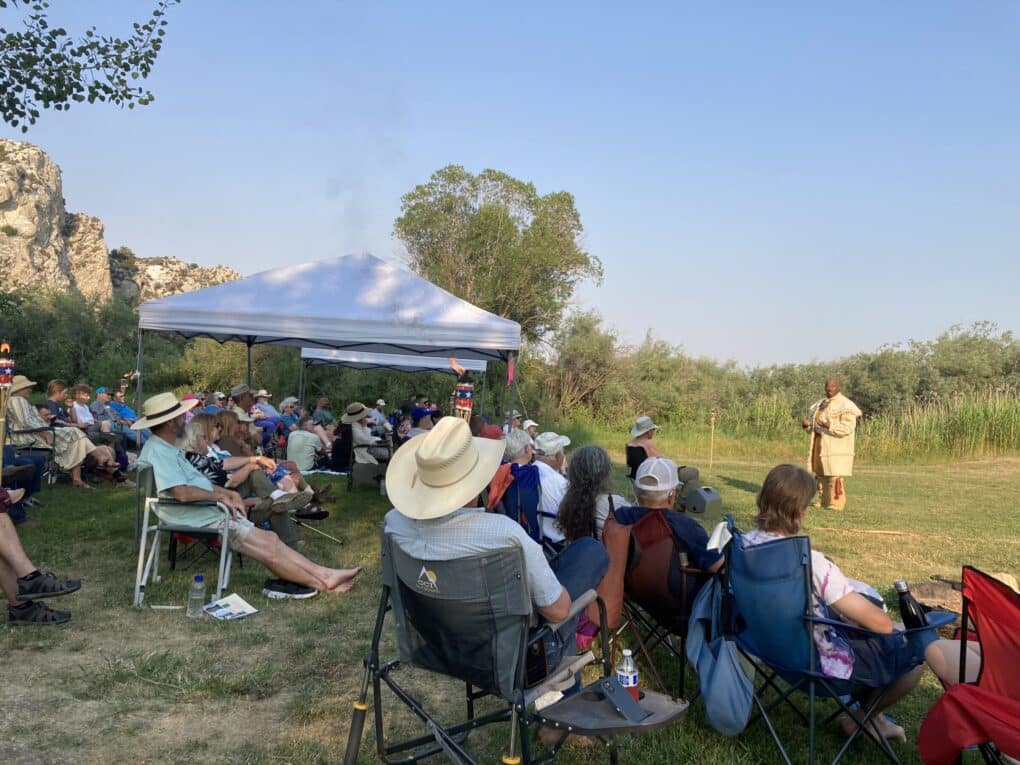 Crowd of people in lawnchairs on a grassy lawn watching a man speak. The man is black, bald, tall, broad-shouldered, wearing what appears to be a buckskin coat.
