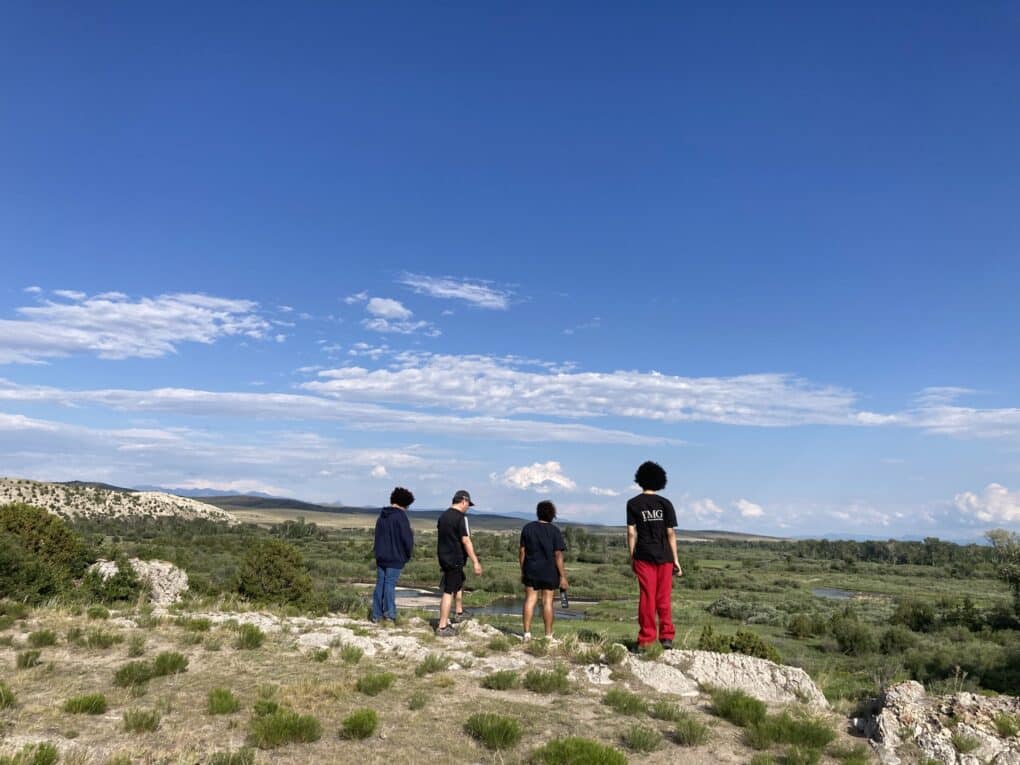 Landscape photo of low hills with sparse vegetation, blue sky, and 4 people at some distance from the camera, facing away. 
