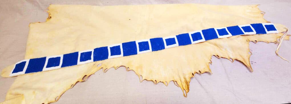 A beaded blue belt is laying on an tanned animal hide. The belt has a repeating pattern of solid blue squares and white and blue squares. 