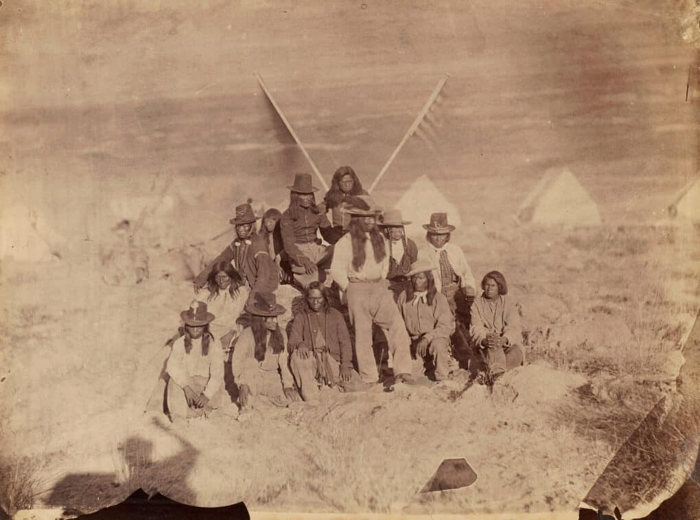 Very old sepia-tone group photo of Shoshone. Many are wearing broad brim hats, with long hair. 