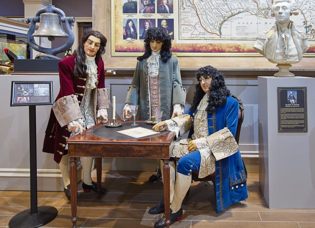 Three wax figures around a table, wearing wigs and colorful 18th-century clothing. 