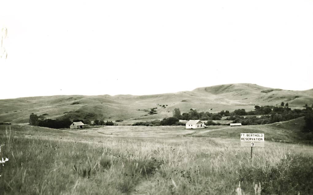 Old black and white landscape photo of hills and a few scattered houses with small sign, "Ft. Berthold Reservation."
