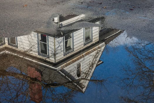 Color photograph of clapboard house reflected in a pool of water in the street, with a white picket fence (the house across the street) at top of image.