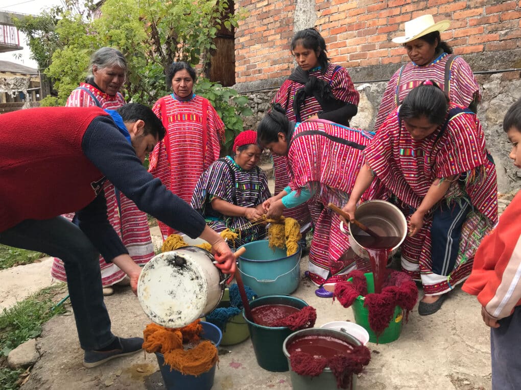 Color photograph of eight people, six women, one little boy, and one man in a paved space beside a brick building, pouring dyes from pots into buckets.