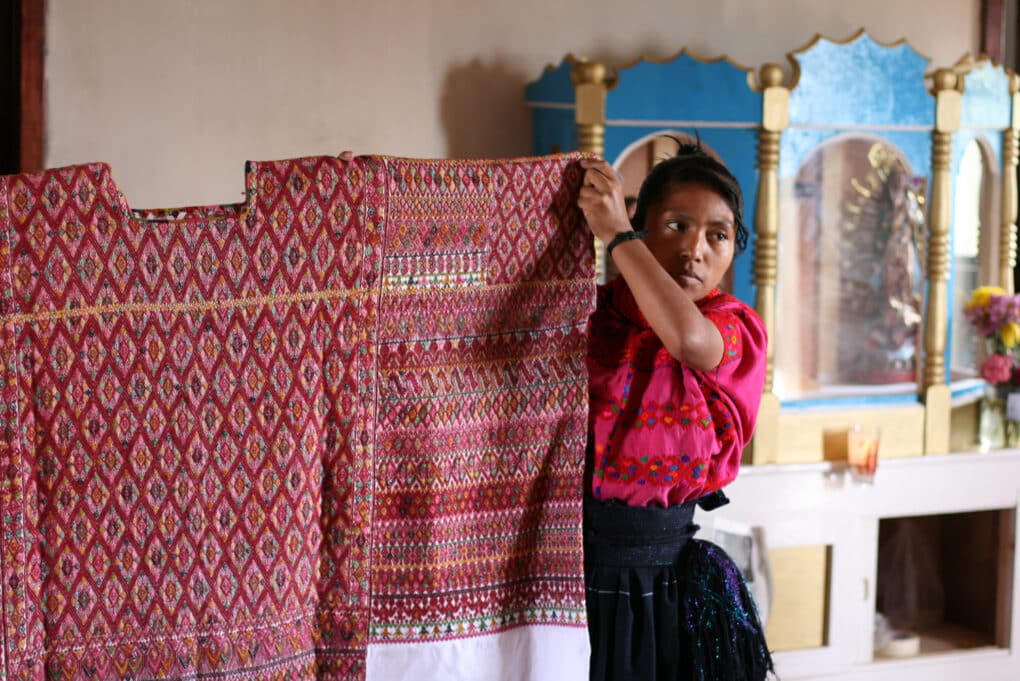 Color photograph of young woman in pink blouse holding up an elaborately woven Mayan huipil.