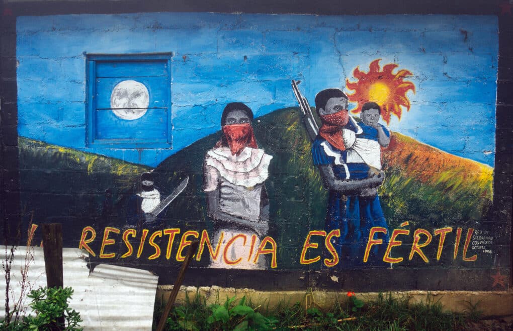 Color photograph of mural showing masked man and woman, the man holding a child and a gun. Behind them, a fiery sun rises over a green mountain. In the foreground, large letters read RESISTENCIA ES FERTIL.
