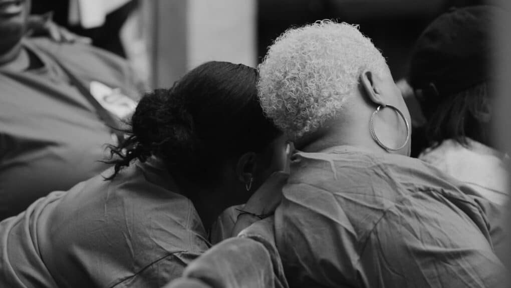 Black-and-white photograph of two Black women, seen from the back, with their heads leaning together.