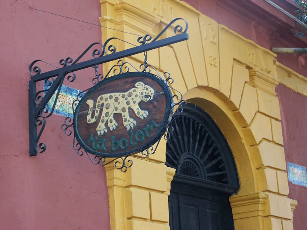 Color photograph of dark-purple stucco wall and yellow arched entrance, with a hanging wooden sign with a figure of a jaguar and the words "Na Bolom."