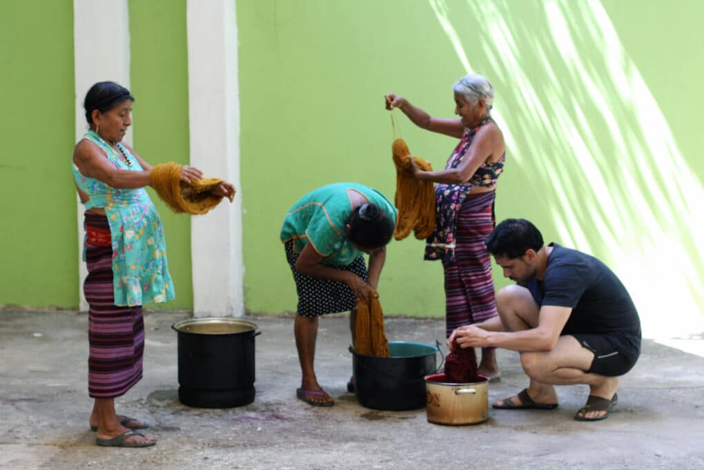 Color photograph of three women and one man dyeing skeins of wool in front of a bright green-painted wall.