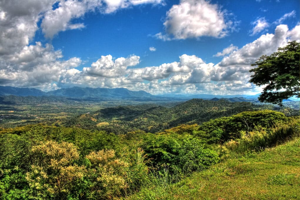 Color photograph of mountainous green country under blue sky with white clouds.