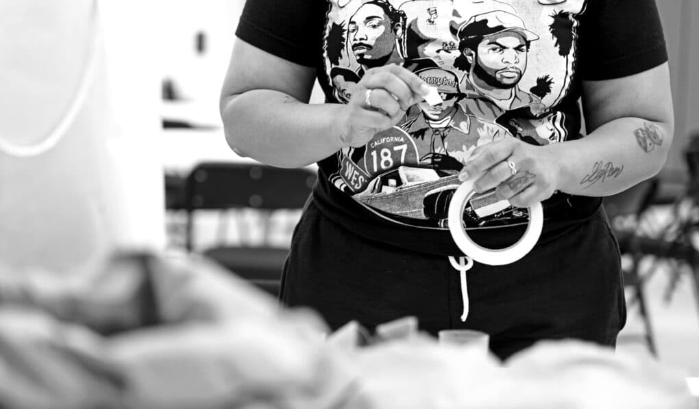 Black-and-white photograph of a young black woman's hands holding a roll of white artist's tape. She is wearing a black t-shirt with white drawings of the faces of west-coast rappers and a road sign for California 187.